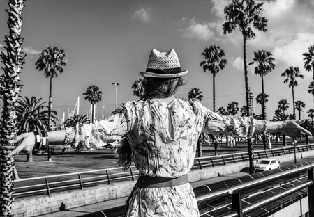 Summertime at colorful Barcelona. Seen from behind elegant tourist woman in long dress and straw hat on embankment in Barcelona, Spain rejoicingの写真素材