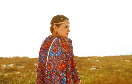 Bohemian vibe vacation. Seen from behind modern gypsy style girl in jeans shorts and cape outdoors in the summer evening looking into the distanceの写真素材
