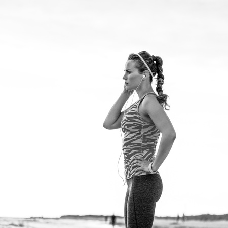 Full length portrait of pensive fit woman in sport clothes on the seashore looking into the distance and listening to the music with headphonesの写真素材