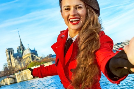 happy young traveller woman in red trench coat on embankment in Paris, France holding friends hand and pointing at Notre Dame de Parisの写真素材