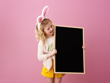 Festive bunny and eggs season. smiling stylish girl in Easter bunny ears on pink background looking at blank boardの写真素材