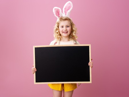 Festive bunny and eggs season. smiling trendy child in Easter bunny ears isolated on pink background showing blank boardの写真素材