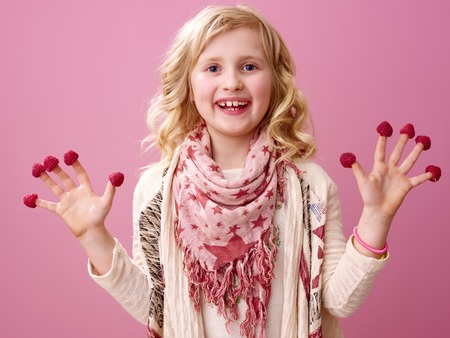 Pink mood. Portrait of happy stylish girl with wavy blonde hair on pink background showing raspberries on the fingersの写真素材