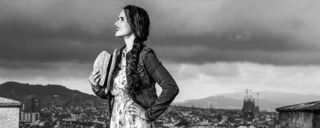 Perfect evening with stunning view. happy elegant traveller woman in long dress and straw hat in the front of city panorama of Barcelona, Spain looking into the distanceの写真素材