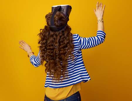 Seen from behind trendy woman in striped jacket on yellow background using VR headset
