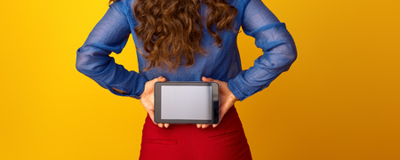 Seen from behind stylish woman with long wavy brunette hair showing tablet PC blank screen on yellow backgroundの写真素材