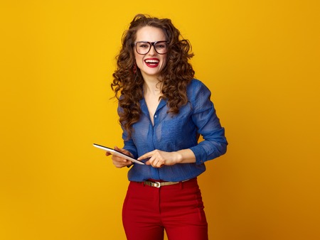 smiling modern woman with long wavy brunette hair using tablet PC against yellow backgroundの写真素材