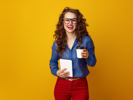 smiling stylish woman with long wavy brunette hair with a coffee cup and tablet PC against yellow backgroundの写真素材