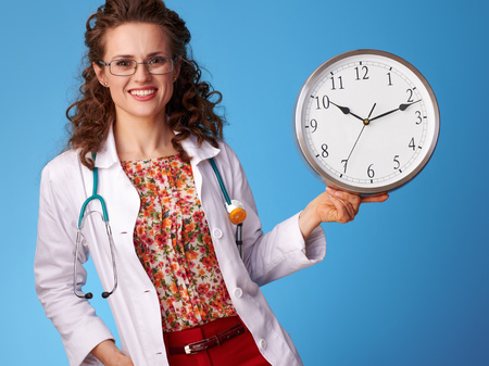 smiling paediatrician doctor in white medical robe showing clock isolated on blueの写真素材