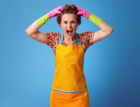 Big cleaning time. Portrait of stressed modern woman in a yellow apron isolated on blueの写真素材