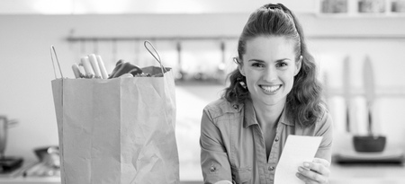 Smiling young housewife examines purchases and check after shopping in kitchenの写真素材