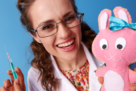 smiling paediatrist doctor in white medical robe with syringe distracting child with toy isolated on blue backgroundの写真素材
