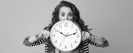 surprised modern woman with long wavy brunette hair isolated on background hiding behind clockの写真素材