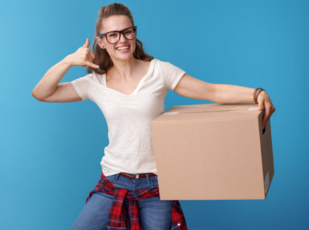 smiling young woman in white shirt with cardboard box showing call me gesture isolated on blue...