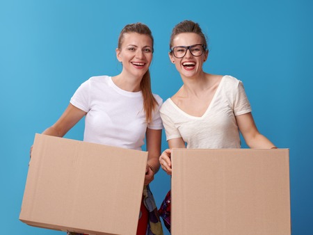 Portrait of happy young female friends in white shirts holding a cardboard boxes against blue backgroundの写真素材