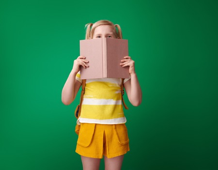 school girl with backpack hiding behind an open book on green backgroundの写真素材