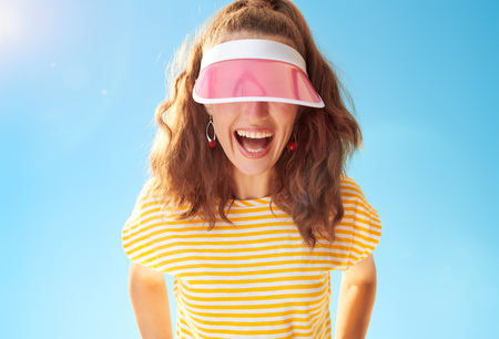cheerful young woman in yellow shirt against blue sky hiding behind sun visorの写真素材