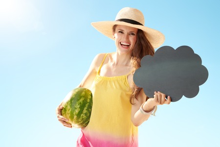 Happy fit woman in straw hat with watermelon showing blank cloud board against blue skyの写真素材