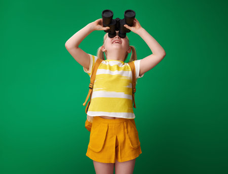smiling school girl with backpack looking up through binoculars isolated on green