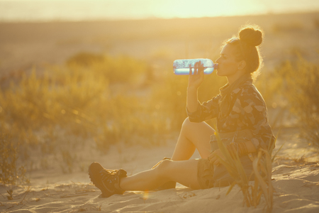 adventure traveller woman in hiking clothes on the seacoast at sunset drinking water from bottleの写真素材