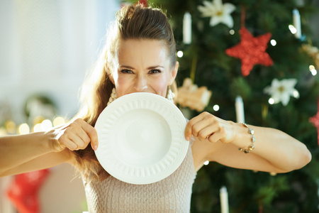 happy modern housewife near Christmas tree hiding behind serving white dinner plateの写真素材