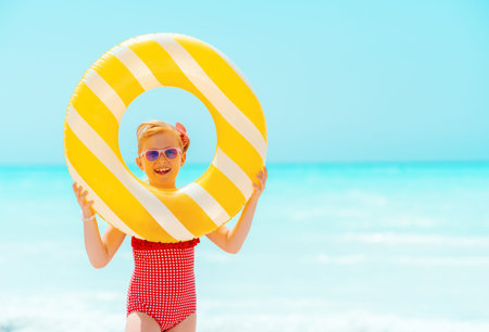 Portrait of happy modern child in red swimwear with yellow inflatable lifebuoy on the seashore