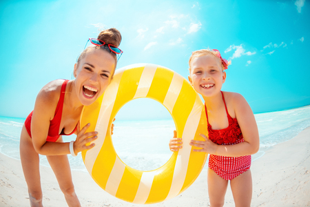 Portrait of happy modern mother and daughter in red swimwear on the seacoast holding yellow inflatable lifebuoyの写真素材