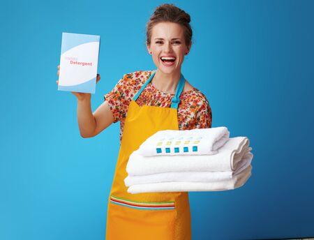 smiling young woman in orange apron with white linen showing powder detergent isolated on blue background. Effective washing powder - a woman shows a detergent to which she was satisfiedの写真素材