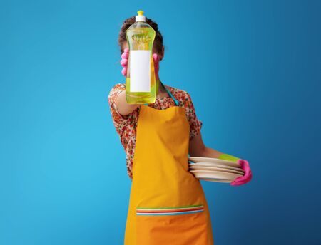 young housewife in orange apron with washed plates showing dishwashing detergent on blue background. housewife advertising effective dishwashing detergent in a large dispenser bottle with blank labelの写真素材
