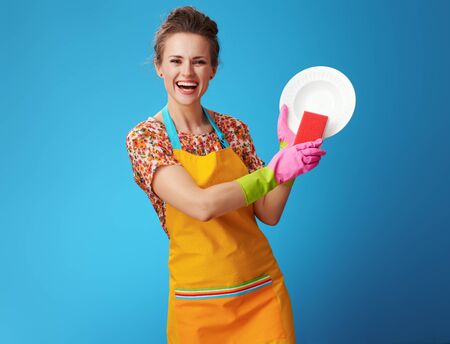 happy young woman in orange apron with sponge washing dish against blue background. Everyone has their own way to relax. You can perfectly meditate at the time of washing up.の写真素材