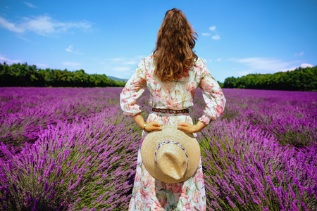 Seen from behind elegant woman in floral dress with straw hat against lavender field of Provence, France. woman enjoying irresistible charm of France during walk in the fresh air of rural siteの写真素材
