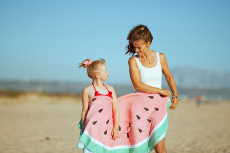 modern mother and child in swimwear wrapped in funny watermelon towel on the seashore in the evening looking at each other. blond hair child in red dotted swimsuit with flowers. modern european motherの写真素材