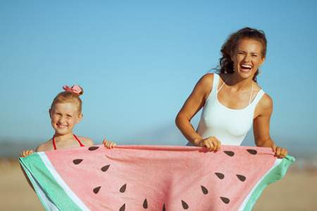 smiling young mother and daughter in swimsuit on the seashore in the evening showing funny watermelon towel. fun and active games for kids to play on the beach. blue sky. modern Ñaucasian mother.の写真素材