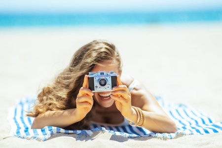 Closeup on retro photo camera holding by woman laying on a striped towel on the ocean shore. Capture...