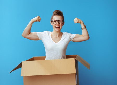 happy young woman in white t-shirt showing biceps in a cardboard box isolated on blue backgroundの写真素材