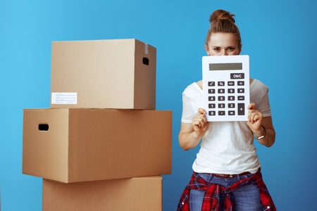 modern woman in white t-shirt near cardboard box hiding behind calculator isolated on blue background. Moving on budget.