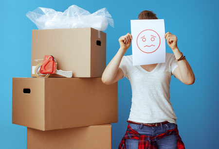 modern woman in white t-shirt near cardboard box hiding behind a sheet of paper with unhappy smiley isolated on blueの写真素材