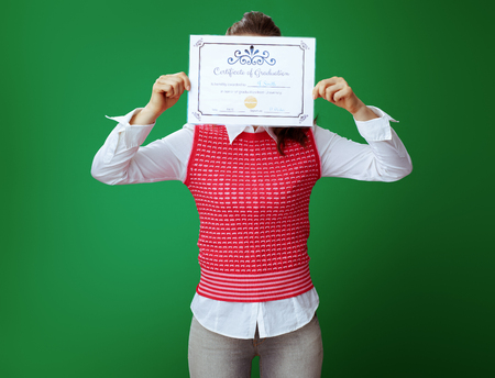 healthy learner woman in grey jeans and pink sleeveless shirt holding Certificate of Graduation in the front of face against green background.の写真素材