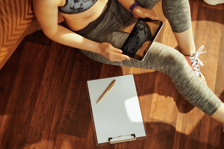 Closeup on healthy sports woman in fitness clothes with clipboard and pen ready to take notes while using online fitness training program in tablet PC at modern home.の写真素材