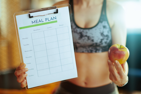 Closeup on young woman in sport clothes showing an apple and clipboard with meal plan at modern home.の写真素材