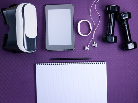 Closeup on VR glasses, white fitness tracker, headphones, tablet PC, black dumbbells and opened notebook with black pen laying on purple fitness mat. Flat Lay.の写真素材