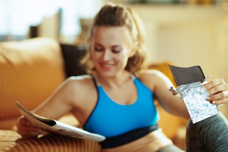 Closeup on chocolate bar in hands of fit woman with magazine in the modern living room.の写真素材