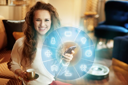 happy young woman with long brunette hair with coffee cup using smart home application on smartphone and robot vacuum cleaning floor in background in the modern living room.の写真素材