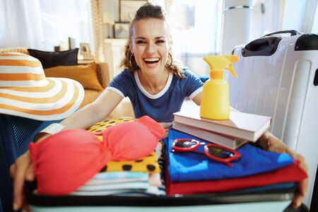 smiling modern woman in blue t-shirt with open suitcase ready for resort in the modern living room in sunny summer day.の写真素材