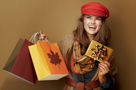 smiling middle age woman in red cap, scarf, jeans shirt and cardigan with autumn shopping bags and golden gift card isolated on beige.の写真素材