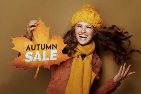 cheerful modern woman in yellow beret and scarf showing autumn sale banner isolated on brown...