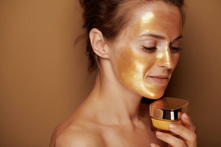 Portrait of pensive modern woman with golden cosmetic face mask holding cosmetic product jar on brown background.の写真素材