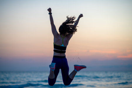 Silhouette of fit woman in sport style clothes with raised arms rejoicing on the seashore in the...