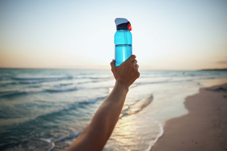 Closeup on famale hand showing bottle of water on the beach at sunset.
