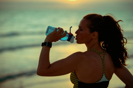 Silhouette of young sports woman in sport style clothes on the ocean shore at sunset drinking water...
